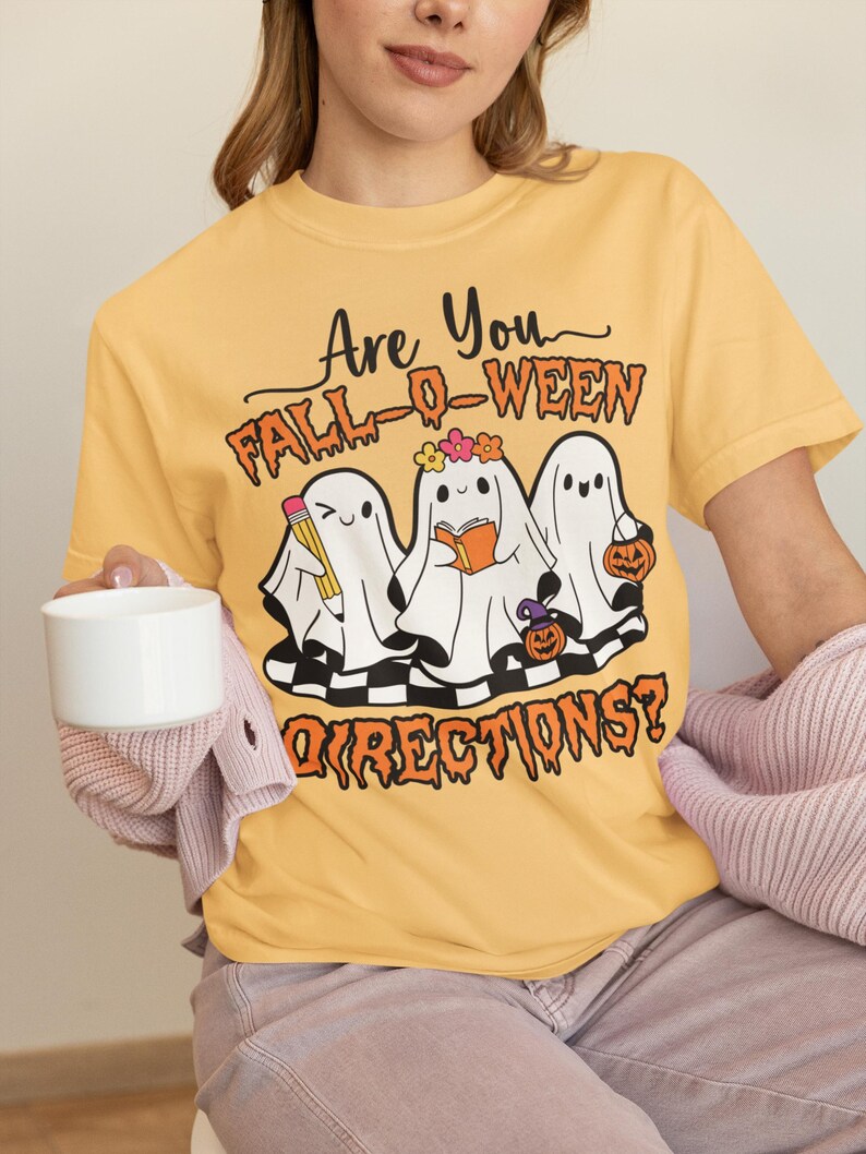 K&ouml;nnte beinhalten: Senfgelbes T-Shirt mit dem Text "Are You Fall-O-Ween Directions?" und einer Grafik von drei Cartoon-Geistern. Ein Geist h&auml;lt einen Bleistift, ein anderer ein Buch und der dritte einen K&uuml;rbis. Die Geister befinden sich auf einem schwarz-wei&szlig;en Schachbrettmuster.
