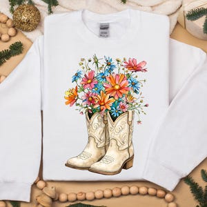 May include: White sweatshirt with a vibrant floral design. The graphic shows cream-coloured cowboy boots filled with a bouquet of pink, orange, blue, and yellow flowers. The boots have decorative stitching and a brown heel.