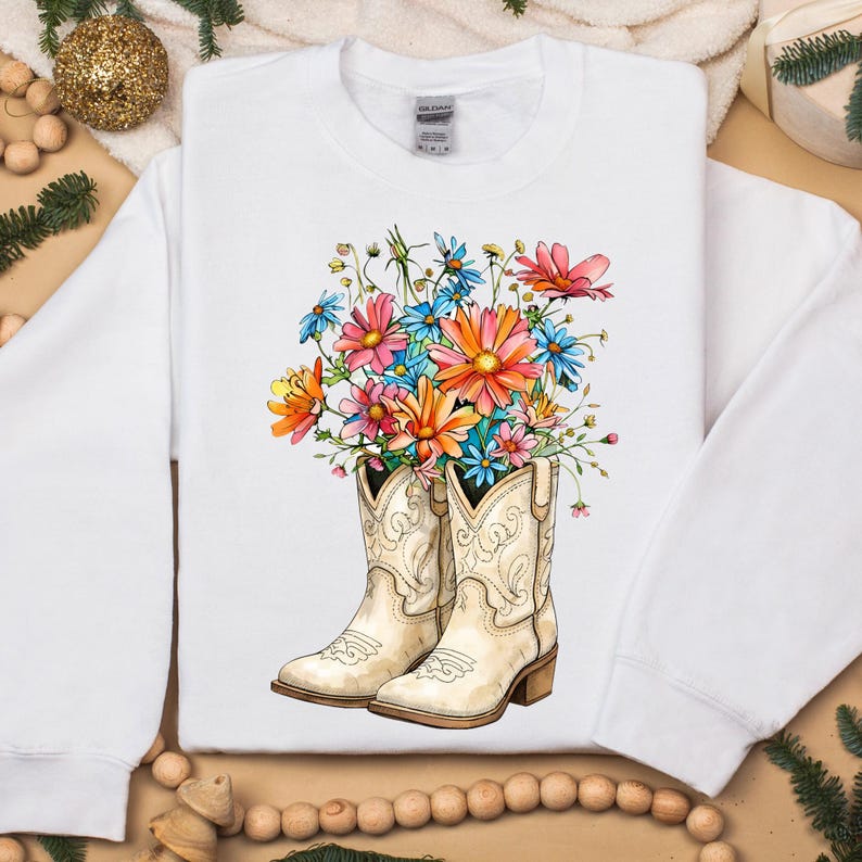 May include: White sweatshirt with a vibrant floral design. The graphic shows cream-coloured cowboy boots filled with a bouquet of pink, orange, blue, and yellow flowers. The boots have decorative stitching and a brown heel.