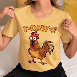 May include: Mustard yellow t-shirt with the text "F-CAW-F" above a cartoon rooster illustration. The rooster is brown with a red comb and is depicted with an open beak. The shirt has short sleeves and a crew neck.