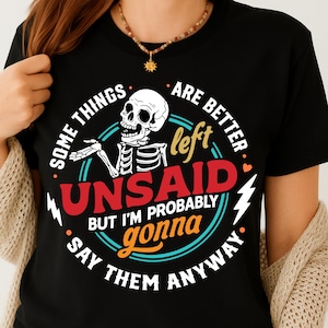 Puede incluir: Camiseta negra con un diseño gráfico circular que presenta un esqueleto y el texto "SOME THINGS ARE BETTER LEFT UNSAID BUT I'M PROBABLY GONNA SAY THEM ANYWAY." El diseño incluye detalles en rojo, blanco, amarillo y turquesa. Una camiseta divertida e informal.