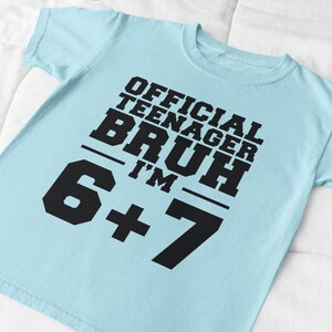 May include: A light blue t-shirt with the text "OFFICIAL TEENAGER BRUH I'M 6+7" in bold black letters. The shirt is made of a soft material and is suitable for casual wear.