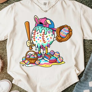 May include: White t-shirt featuring a whimsical baseball-themed Easter design. The graphic includes a baseball character with a bat, glove, and Easter eggs, along with a basket of eggs and colorful sneakers. The design incorporates pastel colors.