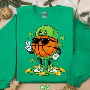 Op de afbeelding: Groen sweatshirt met een cartoon basketbal met een zonnebril en een groene pet versierd met klavers. Het basketbalpersonage maakt een vredesteken en is omringd door gouden munten en klavers. Het woord "PNG" staat onderaan.
