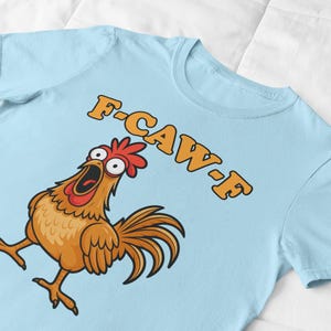 May include: Light blue t-shirt featuring a cartoon chicken with an open beak and the text "F-CAW-F" in orange. The chicken is brown, with a red comb and tail feathers. The shirt is made of a soft material.