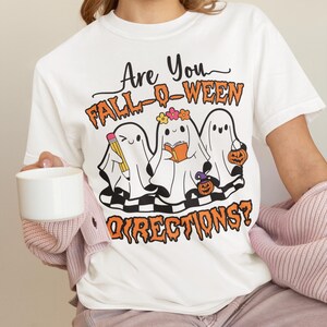 K&ouml;nnte beinhalten: Wei&szlig;es T-Shirt mit einem Halloween-Motiv. Das Design zeigt drei Cartoon-Geister mit Accessoires, den Text "Are You Fall-O-Ween Directions?" in Orange und Schwarz und ein Schachbrettmuster.
