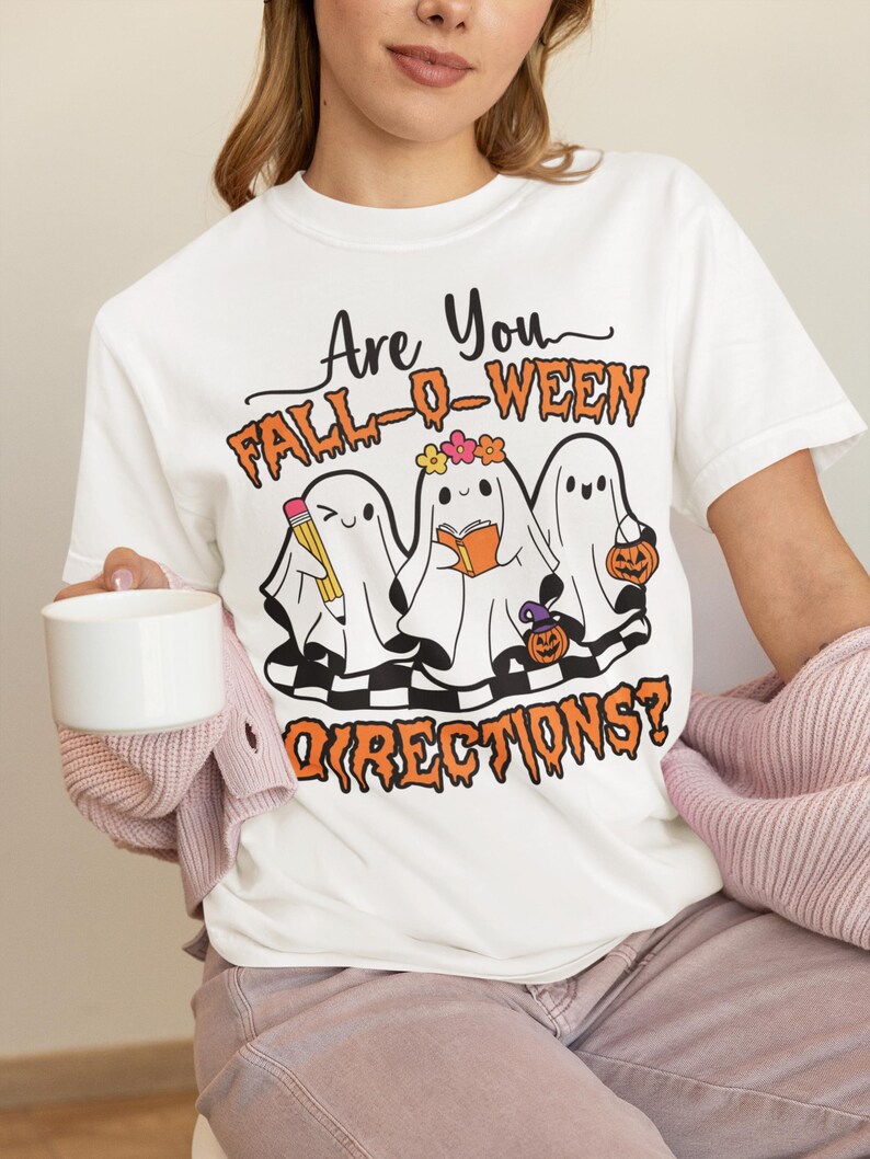 K&ouml;nnte beinhalten: Wei&szlig;es T-Shirt mit einem Halloween-Motiv. Das Design zeigt drei Cartoon-Geister mit Accessoires, den Text "Are You Fall-O-Ween Directions?" in Orange und Schwarz und ein Schachbrettmuster.