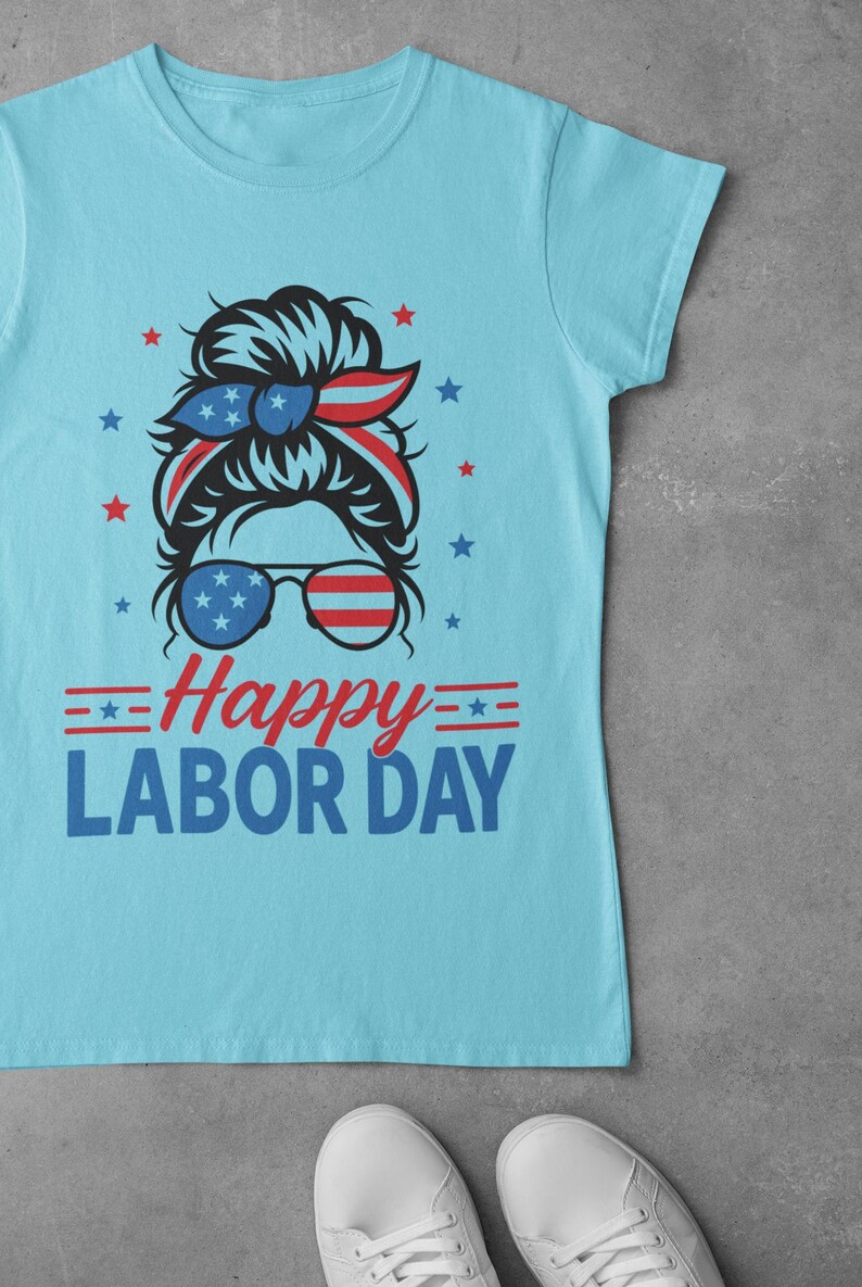 May include: Light blue t-shirt with a graphic design for Labor Day. The design features a woman's face with sunglasses and a headband in red, white, and blue, along with the text "Happy Labor Day."