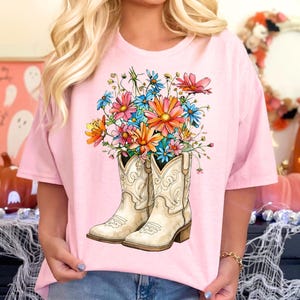 May include: Pale pink t-shirt with a graphic of white cowboy boots filled with colourful flowers. The flowers are in shades of pink, orange, blue, and yellow. The boots have decorative stitching.