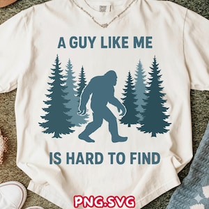 May include: White t-shirt with the text "A Guy Like Me Is Hard To Find" in blue. The design features a silhouette of Bigfoot walking in front of stylized blue pine trees. The shirt is made of a soft material.