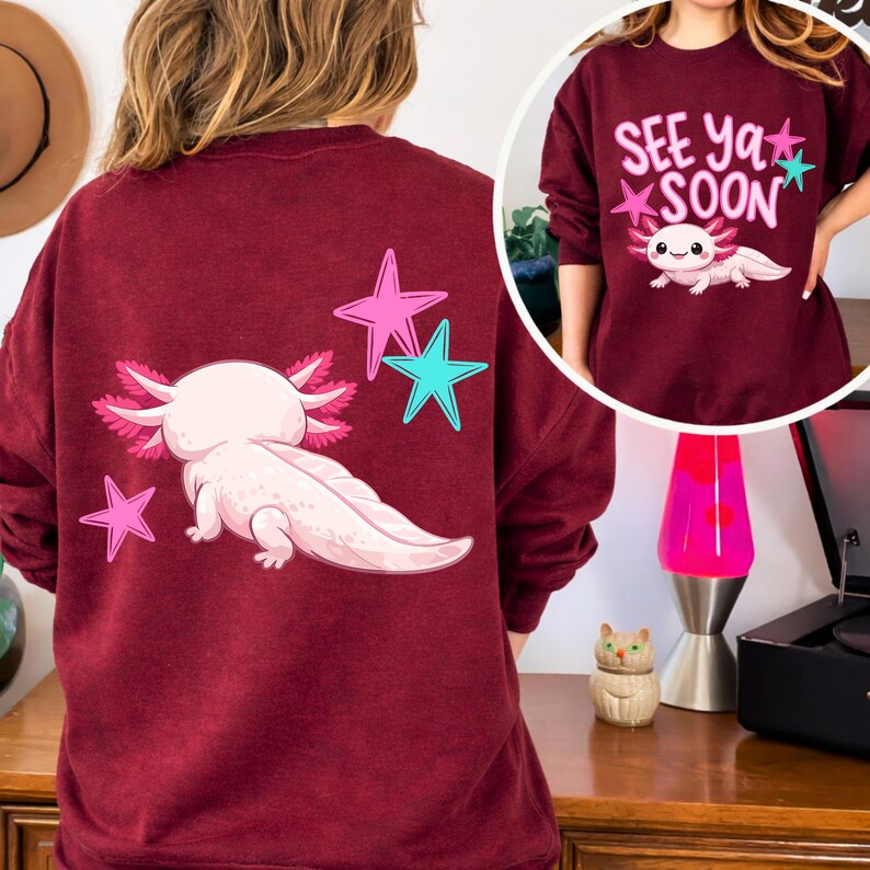 Pu&ograve; includere: Felpa bordeaux con un axolotl dei cartoni animati e stelle. La parte posteriore della felpa mostra l'axolotl e tre stelle. La parte anteriore della felpa ha il testo "See Ya Soon" e un axolotl.