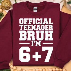 May include: A maroon sweatshirt with the text "OFFICIAL TEENAGER BRUH I'M 6+7" in white block letters. The sweatshirt has a crew neck and long sleeves. The Gildan brand tag is visible on the back of the neck.