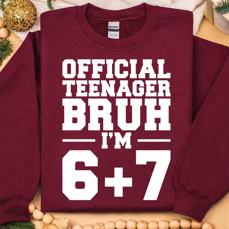 May include: A maroon sweatshirt with the text "OFFICIAL TEENAGER BRUH I'M 6+7" in white block letters. The sweatshirt has a crew neck and long sleeves. The Gildan brand tag is visible on the back of the neck.