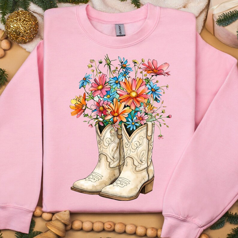 May include: Pale pink sweatshirt with a floral design. The graphic shows white cowboy boots filled with a colourful bouquet of flowers in shades of pink, orange, blue, and yellow. The sweatshirt has a crew neck and long sleeves.