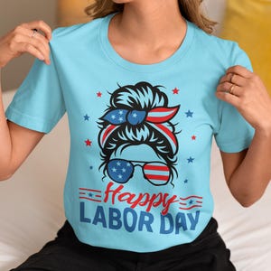 May include: Light blue t-shirt with a graphic celebrating Labor Day. The design features a woman's face with sunglasses and a headband in red, white, and blue, along with the words "Happy Labor Day".
