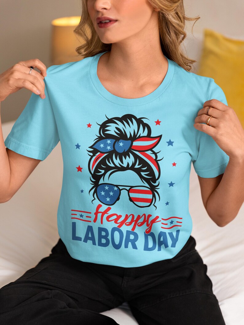 May include: Light blue t-shirt with a graphic celebrating Labor Day. The design features a woman's face with sunglasses and a headband in red, white, and blue, along with the words "Happy Labor Day".