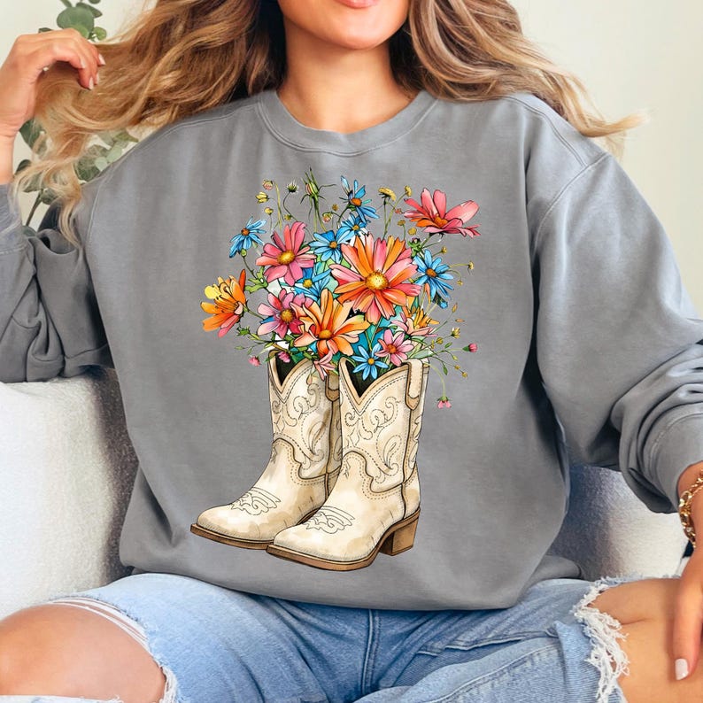 May include: A grey sweatshirt with a watercolour design of white cowboy boots filled with colourful flowers. The flowers include shades of pink, orange, blue, and yellow. The boots have decorative stitching and a small heel.
