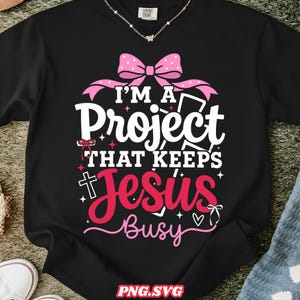 Peut inclure: T-shirt noir à manches longues avec l'inscription "I'M A Project That Keeps Jesus Busy" en blanc et rose. Le motif comprend un nœud rose, une croix et des étoiles. Le t-shirt est en matière souple.
