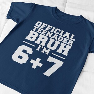 May include: A navy blue t-shirt with the text "OFFICIAL TEENAGER BRUH I'M 6+7" in white block letters. The shirt is made of a soft material and is suitable for casual wear. The text is centered on the front of the shirt.