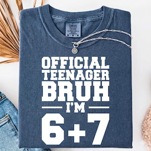 May include: A navy blue t-shirt with the text "OFFICIAL TEENAGER BRUH I'M 6+7" in white block letters. The shirt is folded and has a silver necklace with a small pendant. The shirt is made of Comfort Colors material.