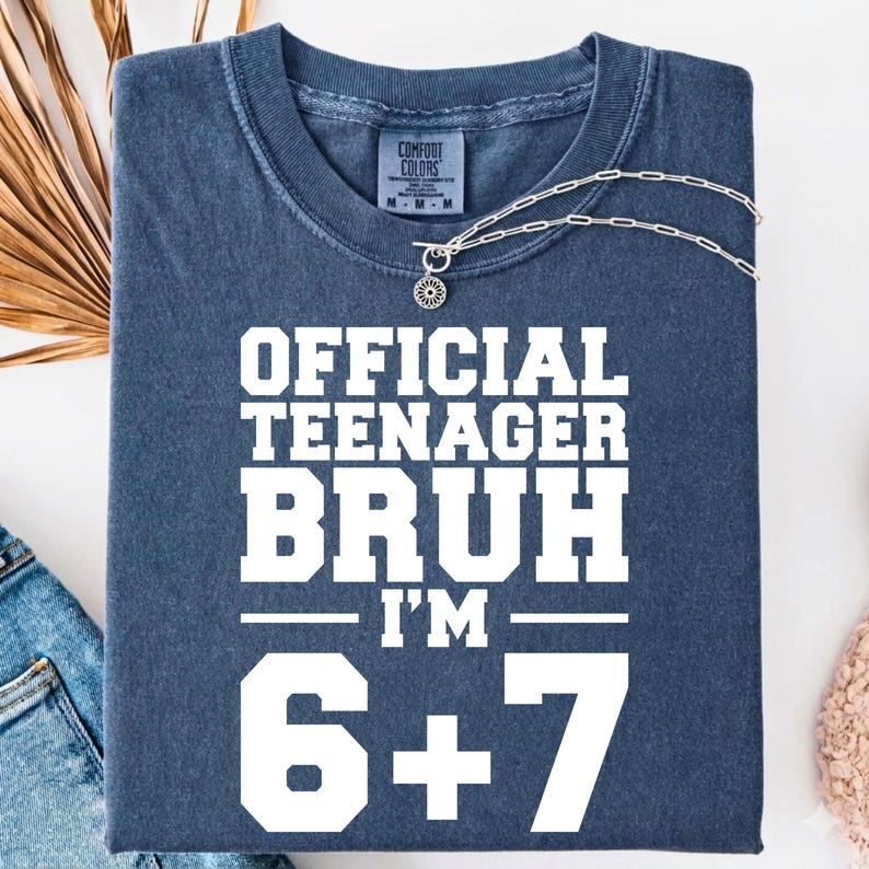 May include: A navy blue t-shirt with the text "OFFICIAL TEENAGER BRUH I'M 6+7" in white block letters. The shirt is folded and has a silver necklace with a small pendant. The shirt is made of Comfort Colors material.