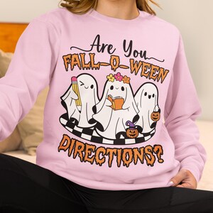 K&ouml;nnte beinhalten: Hellrosa Sweatshirt mit dem Text "Are You Fall-O-Ween Directions?". Die Grafik zeigt drei Cartoon-Geister mit einem Bleistift, einem Buch und einem K&uuml;rbis. Die Geister stehen auf einer schwarz-wei&szlig; karierten Plattform.