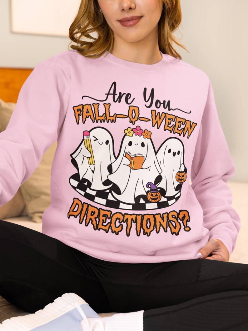 K&ouml;nnte beinhalten: Hellrosa Sweatshirt mit dem Text "Are You Fall-O-Ween Directions?". Die Grafik zeigt drei Cartoon-Geister mit einem Bleistift, einem Buch und einem K&uuml;rbis. Die Geister stehen auf einer schwarz-wei&szlig; karierten Plattform.