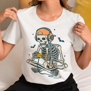 May include: White t-shirt featuring a skeleton wearing an orange cap and headphones, playing a video game. The skeleton is seated on a skateboard, with bats and stars in the background. The design is in shades of blue, orange, and yellow.