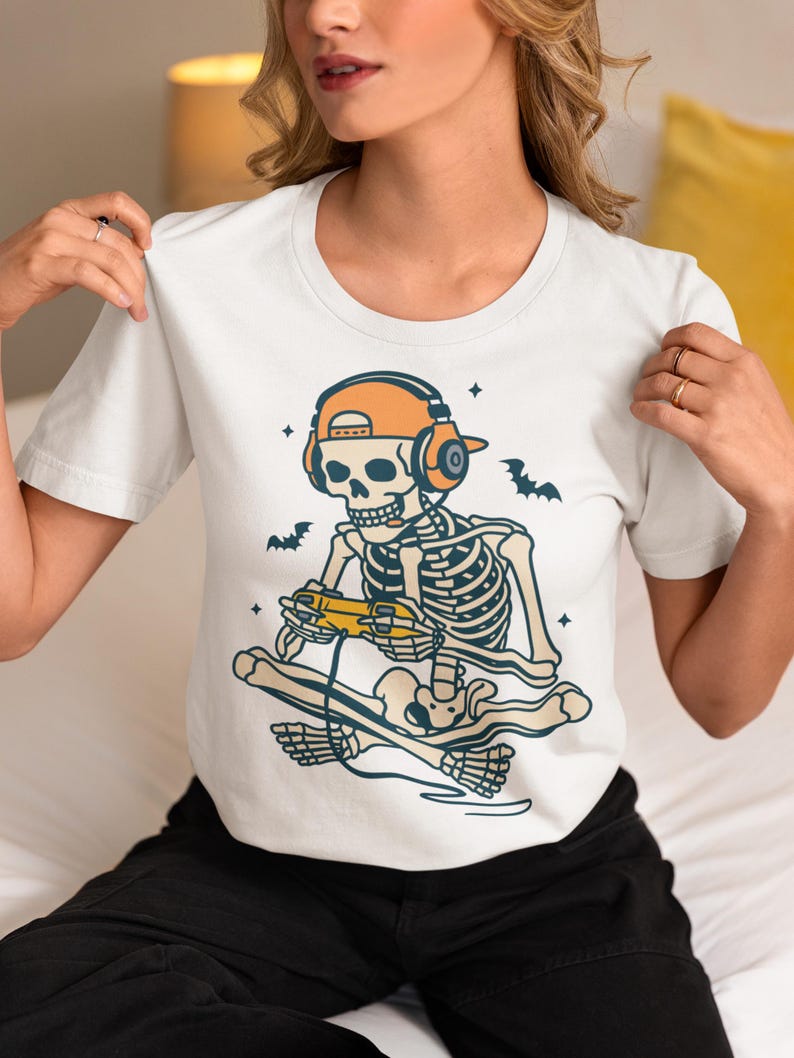 May include: White t-shirt featuring a skeleton wearing an orange cap and headphones, playing a video game. The skeleton is seated on a skateboard, with bats and stars in the background. The design is in shades of blue, orange, and yellow.