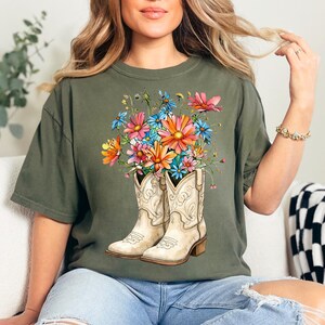 May include: Olive green t-shirt with a graphic of white cowboy boots filled with colourful flowers. The flowers are pink, orange, blue, and yellow. The boots have decorative stitching and a brown heel.