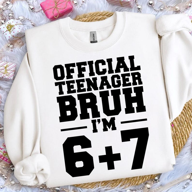 May include: White crewneck sweatshirt with the text "OFFICIAL TEENAGER BRUH I'M 6+7" in bold black letters. The sweatshirt has a classic, casual design, suitable for everyday wear. The text is centered on the front.