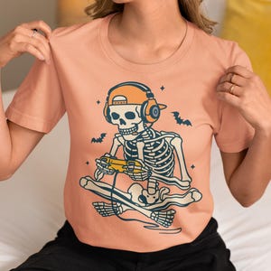 May include: A peach-colored t-shirt featuring a skeleton gamer design. The skeleton wears headphones and a cap, holding a game controller while sitting on a skateboard. Bats and stars are also included in the design.