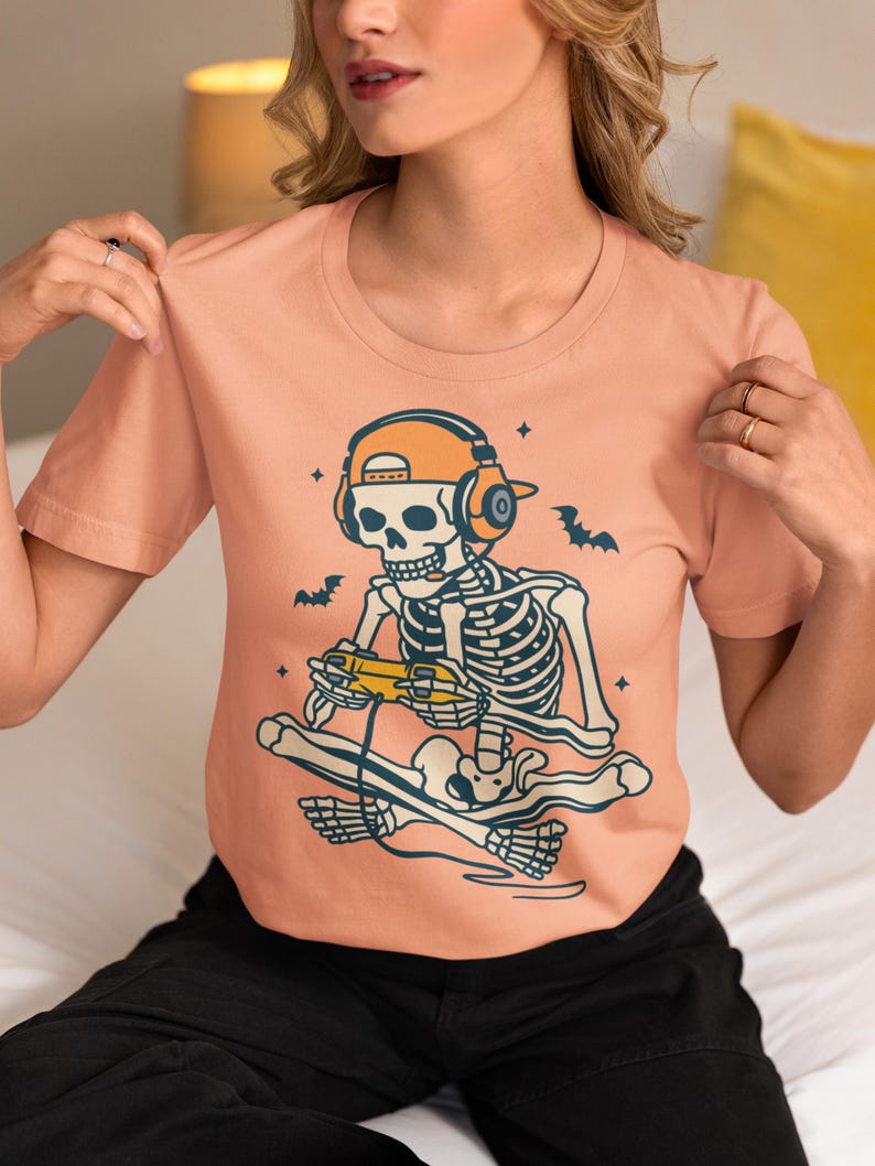 May include: A peach-colored t-shirt featuring a skeleton gamer design. The skeleton wears headphones and a cap, holding a game controller while sitting on a skateboard. Bats and stars are also included in the design.