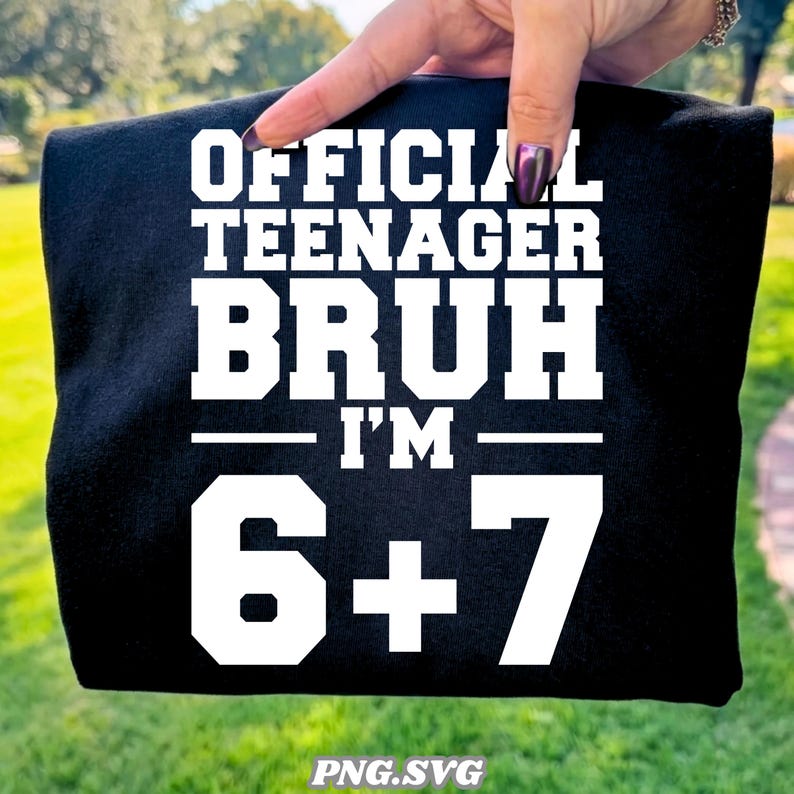 May include: Black t-shirt with white text that reads "OFFICIAL TEENAGER BRUH I'M 6+7". The shirt is held up by a hand against a background of green grass.