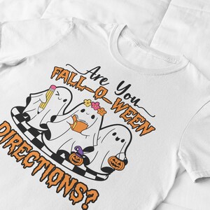 K&ouml;nnte beinhalten: Wei&szlig;es T-Shirt mit einem Halloween-Motiv. Das Design zeigt drei Cartoon-Geister mit Accessoires, ein Schachbrettmuster und den Text "Are You Fall-O-Ween Directions?" in Orange und Schwarz.