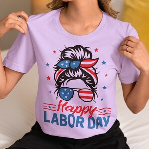 May include: A light purple t-shirt with a graphic design for Labor Day. The design features a woman's face with sunglasses and a headband in red, white, and blue, along with the text "Happy Labor Day".