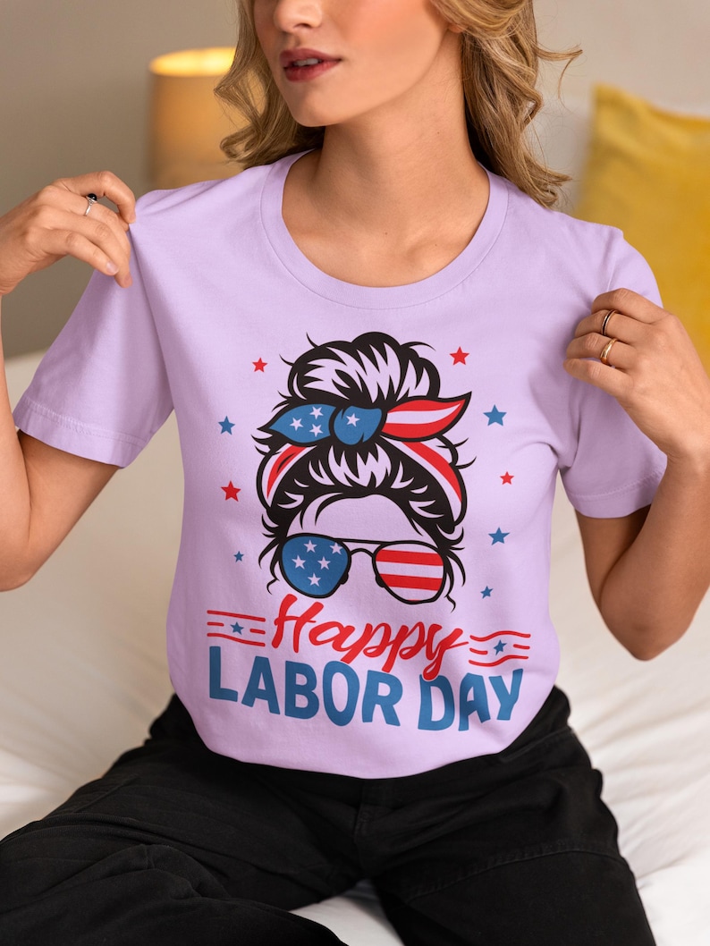 May include: A light purple t-shirt with a graphic design for Labor Day. The design features a woman's face with sunglasses and a headband in red, white, and blue, along with the text "Happy Labor Day".