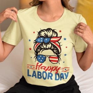 May include: A light yellow t-shirt with a graphic celebrating Labor Day. The graphic features a woman's face with sunglasses and a headband in red, white, and blue. The words "Happy Labor Day" are written in red and blue.