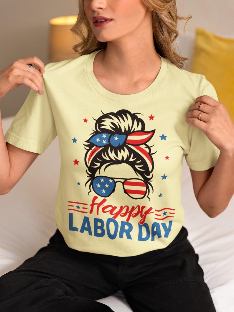 May include: A light yellow t-shirt with a graphic celebrating Labor Day. The graphic features a woman's face with sunglasses and a headband in red, white, and blue. The words "Happy Labor Day" are written in red and blue.