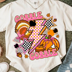 Peut inclure: T-shirt crème avec un motif coloré sur le thème de l'Action de grâce. Le motif comprend une dinde, des citrouilles, des étoiles et le mot "GOBBLE" en rose. Le graphisme a un style rétro et cartoon.