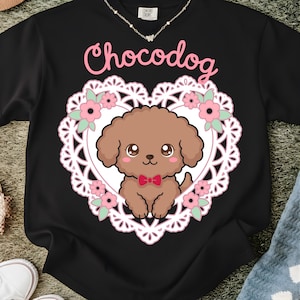 May include: Black sweatshirt featuring a cartoon brown poodle inside a white heart-shaped lace design with pink flowers. The word "Chocodog" is written in pink script above the heart. A delicate necklace is visible at the top.