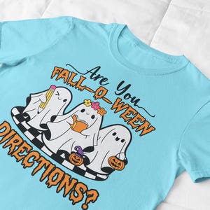 K&ouml;nnte beinhalten: Hellblaues T-Shirt mit einem Halloween-Motiv. Das Design zeigt drei Cartoon-Geister mit Accessoires, ein Schachbrettmuster und den Text "Are You Fall-O-Ween Directions?" in Orange und Schwarz.