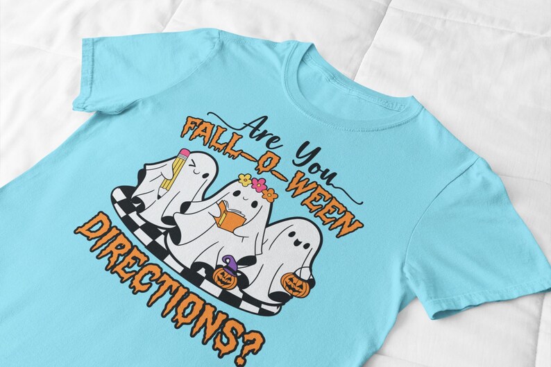 K&ouml;nnte beinhalten: Hellblaues T-Shirt mit einem Halloween-Motiv. Das Design zeigt drei Cartoon-Geister mit Accessoires, ein Schachbrettmuster und den Text "Are You Fall-O-Ween Directions?" in Orange und Schwarz.