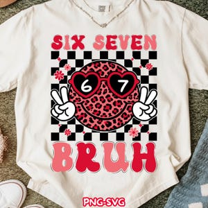 Peut inclure: T-shirt blanc avec un motif graphique. Le motif comprend les mots "SIX SEVEN" en rose, un smiley portant des lunettes de soleil en forme de cœur avec les chiffres 6 et 7, et le mot "BRUH". Le motif comprend des signes de paix et un fond à carreaux.