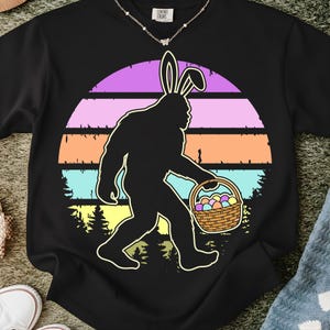 May include: Black t-shirt featuring a Bigfoot silhouette wearing bunny ears and carrying a basket of Easter eggs. The design includes a colorful sunset background with the text "COMFORT COLORS" at the top.