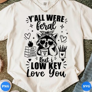 Peut inclure: T-shirt blanc avec un dessin d'un raton laveur portant des lunettes de soleil en forme de cœur et un bandana. Le texte dit "Y'all were feral, but I low key love you." Autres graphiques : cœurs, étoiles et une pile de livres.