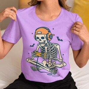 May include: A lilac-colored t-shirt featuring a cartoon skeleton wearing orange headphones and a matching cap, playing a video game while skateboarding. The design includes bats and stars, perfect for Halloween or gaming enthusiasts.