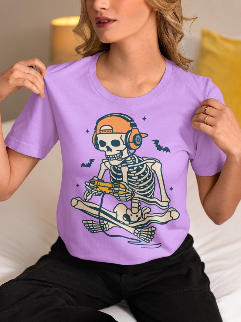 May include: A lilac-colored t-shirt featuring a cartoon skeleton wearing orange headphones and a matching cap, playing a video game while skateboarding. The design includes bats and stars, perfect for Halloween or gaming enthusiasts.