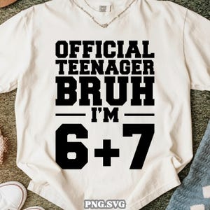 May include: Cream-colored t-shirt with the text "OFFICIAL TEENAGER BRUH I'M 6+7" in bold black letters. The shirt is made of a soft material and is likely for casual wear. The image also shows a small portion of a necklace.