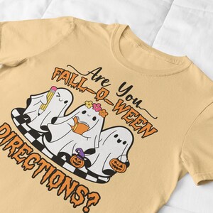 K&ouml;nnte beinhalten: Hellgelbes T-Shirt mit einem Halloween-Motiv. Das Design zeigt drei Cartoon-Geister mit Accessoires und dem Text "Are You Fall-O-Ween Directions?". Die Geister stehen auf einer schwarz-wei&szlig; karierten Plattform.